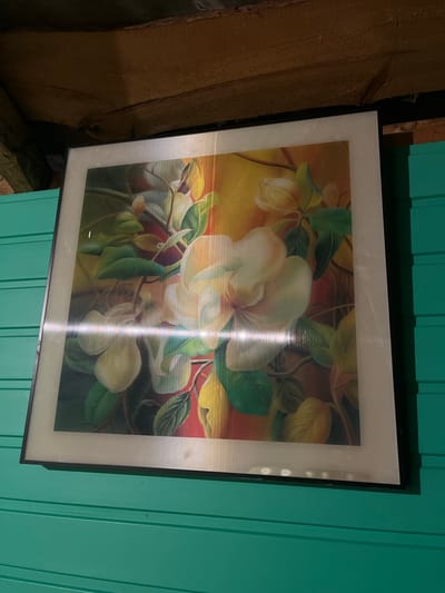 A framed floral painting hangs on a teal-colored, horizontally paneled wall. The painting features large white and light yellow blossoms with green leaves against a background that transitions from yellow at the top to reddish-brown at the bottom. The artwork is framed with a thin black border and a wider white mat. Above the painting, rough-hewn wooden beams form the ceiling, suggesting an interior space. The overall scene is static and depicts a decorative element within a rustic setting. This is a photograph taken in Abzakovo, Russia.