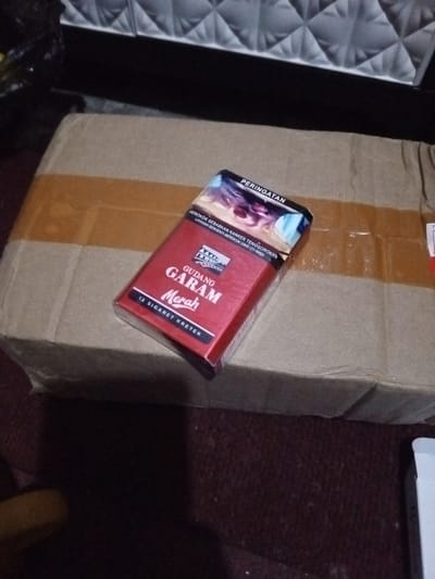 A red pack of Gudang Garam Merah cigarettes rests on a brown cardboard box. The cigarette pack is prominently displayed, featuring the brand name in bold white letters. A health warning regarding throat cancer is printed at the top of the pack. The box underneath appears to be sealed with brown tape. The background includes a quilted headboard and a carpeted surface.