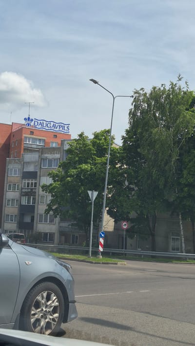 The image captures a view of a building with a prominent sign that reads "DAUGAVPILS" in blue lettering against a light blue background, topped with decorative elements. The building itself appears to be a multi-story structure, with a red brick section at the top and lower levels featuring numerous windows and balconies. Trees with lush green foliage are situated in front of the building and along the roadside, suggesting an urban environment. A street lamp stands tall near the center of the frame. In the foreground, the front portion of a light silver car is visible, indicating the perspective is from inside another vehicle. The scene depicts a sunny day with scattered clouds in the sky. The presence of traffic signs, including one with a truck symbol and another indicating a direction, further confirms an urban street setting. This scene is located in Daugavpils, Latvia.