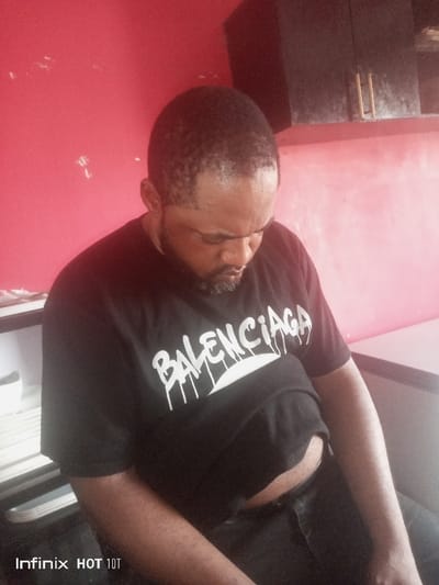 An adult male with dark skin, short dark hair, and a full beard is seated indoors, looking downwards. He is wearing a black short-sleeved t-shirt with "BALENCIAGA" printed in white, stylized, dripping text across the chest, and dark trousers. His midsection is slightly exposed below the t-shirt. The background consists of a predominantly red wall, which shows signs of wear. To the upper right, a dark, wall-mounted cabinet with two horizontal brass-colored handles is visible. To the left, a portion of a shelf or desk with stacked objects is present. The scene is located in a room within the city of Ajaokuta, Nigeria. A watermark "Infinix HOT 10T" is visible in the lower left corner.