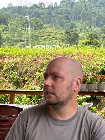 A man is depicted in a close-up, outdoors, in what appears to be Du Già Commune, Vietnam. He is looking downwards and slightly to his right, with a neutral or contemplative expression. The man has a shaved head or very short hair and a short beard or stubble. He is wearing a plain grey t-shirt.

In the immediate foreground, to the man's right, there is a wooden railing and a glimpse of what might be a brown chair or table. Behind him, on an elevated surface, are several white potted plants, some containing small red and orange flowers, suggesting a balcony or porch setting.

The background reveals a lush, verdant landscape of rolling hills or mountains covered in dense green foliage, characteristic of a tropical or subtropical region. Utility lines are visible stretching across the mid-ground, and a white utility pole stands further in the distance against the green backdrop. The lighting suggests it is daytime with clear weather. There is no visible text in the image.