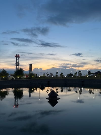 The image captures a serene twilight scene at a park in Novaya Adygeya, Russia. The sky is a canvas of soft blues and oranges, with streaks of gray clouds adding depth. In the distance, industrial structures, including a tall, metal tower with red lights and two smokestacks, punctuate the horizon. The tower and smokestacks are reflected in the still water of a pond in the foreground.
The park itself features a well-maintained embankment lined with trees and lampposts that cast a warm glow on the scene. The water's surface is interrupted by a small, dark, pyramidal structure, likely a duck house or decorative element. The reflection of the sky and the park's features create a mirror image on the water, enhancing the tranquil atmosphere. There are no people visible in the image, emphasizing the quiet and peaceful nature of the park at dusk.