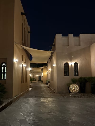 The image shows a night view of a building complex in Doha, Qatar. The buildings are illuminated with warm lighting, highlighting their architectural details, with arched windows. A white canvas awning is suspended above the courtyard area. The walkway is paved with dark grey stone slabs and lined with plants. The night sky is a deep, dark blue.