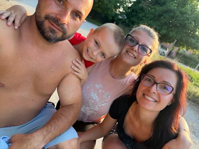 The image is a selfie taken outdoors, likely in the city of Dolni Dabnik, Bulgaria, judging by the presence of deciduous trees and a dirt road. Four individuals are visible: a shirtless man in the foreground on the left, a young boy with his arm around the man's shoulders, a woman wearing glasses and a pink shirt, and another woman wearing glasses and a black shirt. The overall atmosphere suggests a casual, familial gathering during daytime.