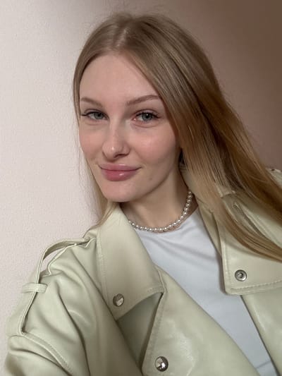 A close-up portrait features a young woman with long, straight, light blonde hair. She has light blue-green eyes and a gentle, pleasant smile, looking directly at the camera. Her lips are slightly glossy. She is wearing a light cream or off-white faux leather jacket, visible over a white t-shirt. A strand of pearls adorns her neck. Details of the jacket, including silver snap buttons and a zipper on the shoulder, are visible. The background is a plain, light beige or off-white surface, suggesting an indoor setting with a simple wall or soft surface. The image itself is rotated counter-clockwise. There is no visible text. The location context provided is Кондратово, Russia.