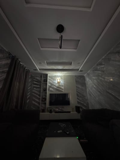 This is an indoor scene that appears to be a modern living room or entertainment space in Kufena, Nigeria. The room is dimly lit, suggesting it's either nighttime or the blinds are drawn to create a cinematic atmosphere.
The primary subjects are the interior design elements. The walls are a mix of smooth, light grey surfaces with decorative rectangular indentations, and a textured, marble-like pattern, particularly on the ceiling and accent walls. One wall features a series of recessed shelves, some of which hold small, dark objects. A prominent object is a black, circular fixture mounted on the wall, connected to a horizontal bar with what looks like a light source or speaker.
A large, dark sofa is visible in the foreground, partially obscuring the view of the rest of the room. To the right, a black surface with a marble pattern is present, holding a remote control and what appears to be a framed picture or tablet. A large, white rectangular object is also on this surface, possibly a television or a piece of furniture. The floor is covered in a dark, glossy material, reflecting the light.
A set of dark, pleated curtains runs along one side of the room, suggesting a window or an entrance is covered. Above the shelves, a mounted light fixture with a textured glass or metal shade casts a warm glow.
The overall impression is one of a sleek, contemporary space designed for relaxation or entertainment. The dim lighting and dark colors contribute to a sophisticated and possibly intimate ambiance. There are no people visible in the image. No discernible text is present.