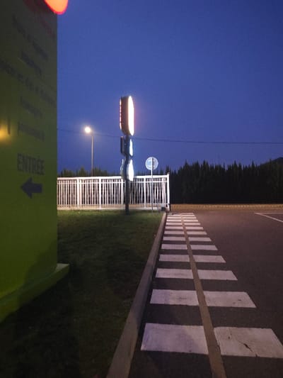 A crosswalk stretches alongside a grassy median, bordered by a low, white fence. Behind the fence stand two tall signs, one glowing with an elongated, rounded rectangular shape and the other circular. A street lamp is visible on the left. Coniferous trees fill the background beneath a dark blue sky. A green building with an arrow pointing left and the word "ENTREE" is on the left edge of the image.