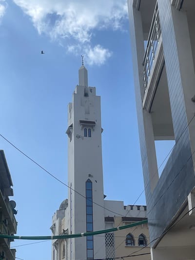 The image displays an upward perspective of the Malindi Mosque's minaret in Stone Town, Zanzibar City, Tanzania, under a clear blue sky with scattered white cumulus clouds. The minaret, a tall, slender, white concrete structure, occupies the central frame. Its apex is a pointed spire crowned with a metallic crescent moon and star finial. Below the spire, four dark loudspeakers are affixed, and multiple rectangular, blue-tinted glass windows extend vertically down one side. Weathering and minor paint chipping are evident on the minaret's white facade. A single dark-colored bird is captured in flight in the upper-left quadrant of the sky. To the right, a portion of a modern, multi-story building with a light grey tiled exterior and structural columns is visible. To the left, an older building, partially obscured, shows elements like air conditioning units and satellite dishes. Several dark utility cables and bundled green cables intersect the view across the lower and middle sections of the image. Structures comprising the mosque's main body and an adjacent yellow-painted building with arched windows are visible in the background. No human subjects are present.