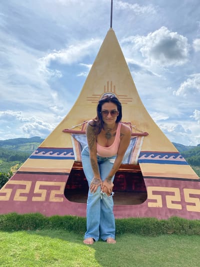 A woman with tattoos and sunglasses is sitting on a colorful, triangular object with indigenous-inspired patterns. She is wearing a pink tank top and light blue jeans. The object appears to be a decorative or artistic installation, possibly a prop. The background shows a bright blue sky with fluffy white clouds and a lush green hillside. The image is taken from a low angle, emphasizing the sky and the structure. The lighting suggests it is daytime, and the weather is clear and pleasant. The overall mood is casual and relaxed. The image is oriented vertically, with the subject positioned in the center and to the left. The grass in the foreground is neatly trimmed.