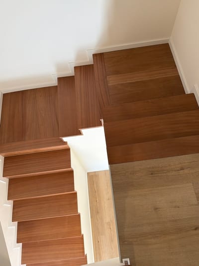 The image shows a top-down view of a modern staircase with dark wooden steps and a lighter wooden floor. The staircase is made of polished dark wood, with a contrasting lighter wood on the landing at the top. The steps are wide and the risers are not visible, creating a sleek, continuous look. A white wall with a minimalist baseboard runs along the side of the stairs. The floor on the landing is a light, natural wood with a visible grain pattern. The environment suggests an interior setting, possibly a contemporary home or building. The image does not contain any people, actions, or interactions. The location in Gondomar, Portugal, is not visually evident from the image itself.