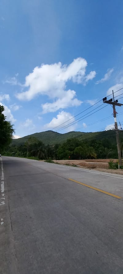 The image shows a road in Baan Chaloklum, Thailand. The road is concrete with a yellow line marking the center. A mountain covered in dense green vegetation rises on one side of the road. Utility poles with power lines run along the edge of the road. The sky is blue with scattered white clouds. Part of what looks like a construction or excavation site is also visible between the road and the mountain vegetation.