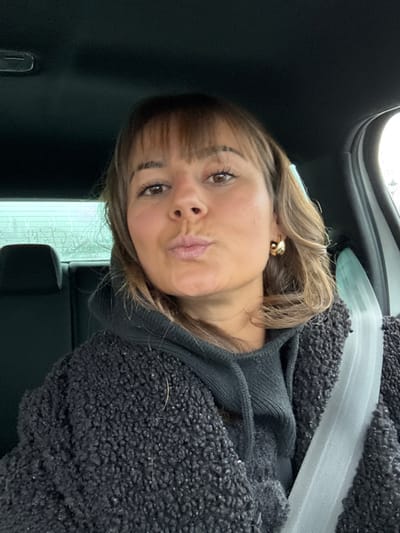 The image depicts a female subject taking a selfie inside a vehicle. The subject is positioned facing the camera, slightly off-center to the left, with her head and torso filling most of the frame. She has medium-length brown hair with bangs, light skin, and brown eyes. Her lips are pursed in a "kiss" expression. She wears a dark grey or black hooded sweatshirt under a textured, dark grey or black jacket, possibly made of a sherpa or bouclé material. A light-colored seatbelt, likely white or light grey, is visible across her left shoulder and chest. A gold-colored hoop earring is visible on her right ear. The vehicle's interior features a dark headliner and dark seatbacks. Through the rear window, behind the subject's left shoulder, blurred tree branches are visible against a light, possibly overcast, sky, suggesting an outdoor environment. The specific city of Saint-Cyprien, France, is not visually verifiable within the image content.