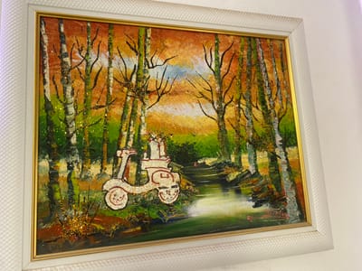 A framed painting hangs on a white wall. The painting depicts a forest scene with birch trees. Two stylized figures, outlined in red, are positioned among the trees, appearing to hold hands. Gold glitter is scattered throughout the painting, particularly on the trees and above them. The painting is framed with a white, textured outer frame and a thin gold inner frame. The background colors are green, brown, orange, and a hint of blue. The style of the painting is naive and abstract.