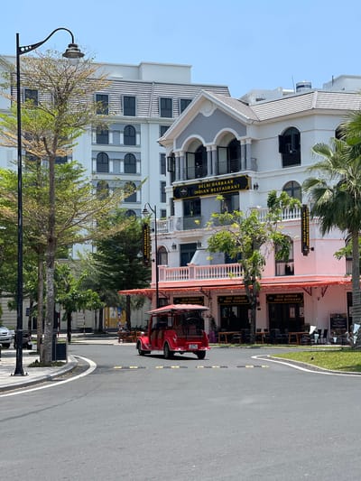 This media file captures a bright, sunny street scene in Phú Quốc, Vietnam. The image is dominated by a multi-story white building with an elegant facade featuring arched windows, balconies with railings, and decorative columns. This building houses "DELHI DARBAAR INDIAN RESTAURANT," with multiple prominent black and gold signs displaying the restaurant's name and cuisine. Orange awnings extend over the ground floor of the restaurant, where outdoor seating with tables and chairs is visible, though no people are present.

The street, made of dark asphalt with white and yellow markings, curves gently to the right. A small, red, open-sided vehicle, resembling a golf cart or tourist shuttle, is parked or driving on the right side of the road. It displays a license plate with the alphanumeric code "51HC 002.71."

Lush green trees and palm trees are interspersed along the street and in front of the buildings, adding a tropical touch to the environment. A black streetlight with an ornate design is visible in the foreground on the left. The sky is clear and blue, indicating bright daylight and good weather. On the rooftop of one of the buildings, a water tank with the letters "XHS" is faintly visible. The overall setting suggests a modern commercial or resort area.