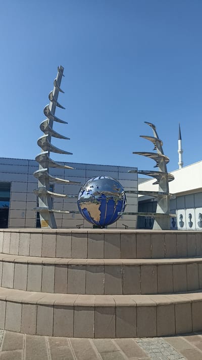 The image depicts an outdoor sculpture featuring a large, reflective globe of the Earth positioned between two abstract, spiraling metal structures. The globe is metallic and shows reflections of the surrounding environment, including parts of the architecture and the sky. The spiraling structures appear to be made of polished metal, with layered, curved elements that create a sense of upward motion.
Behind the sculpture, a modern building with a facade of horizontal metallic panels and a glass section is visible. To the right of the building, a minaret of a mosque pierces the clear blue sky. The foreground is dominated by a tiered, circular stone structure, resembling steps or a fountain base, paved with square stone tiles. The overall scene suggests an urban or public space.
The time of day appears to be daytime, with bright sunlight casting shadows. The sky is a uniform, clear blue, indicating fair weather. There is no visible activity of people, but there are some busts or statues visible on a wall behind the globe on the right side of the building, hinting at a cultural or memorial site.
The location is identified as Yenişehir, Turkey. No significant text is clearly legible in the image, though there may be a plaque at the base of the left spiral structure. The overall mood is serene and somewhat monumental, highlighting the sculpture as a centerpiece within its architectural and environmental context.
