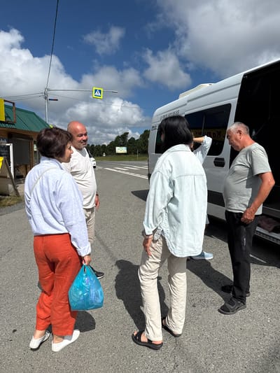 Four people are standing on an asphalt road near a white van with its rear door open. The van has the phone number "8 (383) 581-55-55" printed on its side. A woman in the foreground is wearing orange pants and a light gray sweatshirt, holding a bright blue plastic bag. Beside her stands a bald man in a white t-shirt and light-colored pants. To the right of them, a woman with dark hair wears a light blue denim jacket and beige pants. An older man in a gray t-shirt and black pants stands facing the open van. In the background, a yellow pedestrian crossing sign is visible on a pole, along with a green-roofed building to the left. The sky is blue with scattered white clouds, and trees are visible in the distance. The location is T/B Seminskii Pereval, Russia.