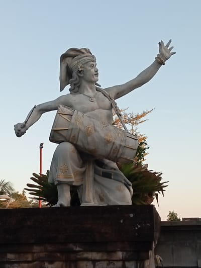 This is a low-angle, outdoor, eye-level shot of a statue of a male figure in Gianyar, Indonesia, against a clear, pale blue sky. The statue, made of light-colored stone, depicts a man in traditional Balinese attire, with a cloth headpiece adorned with a flower. He is seated, playing a large, cylindrical drum that is slung across his chest. His left arm is bent, holding a drumstick, and his right arm is raised, with an open hand reaching upwards. The statue is adorned with gold-colored detailing on the drum and his lower garment. The base of the statue is a dark, textured stone structure, partially obscured by lush green foliage. In the background, a tall red pole with a light fixture is visible, along with trees and other architectural elements suggestive of an urban setting. The lighting indicates it is either early morning or late afternoon.