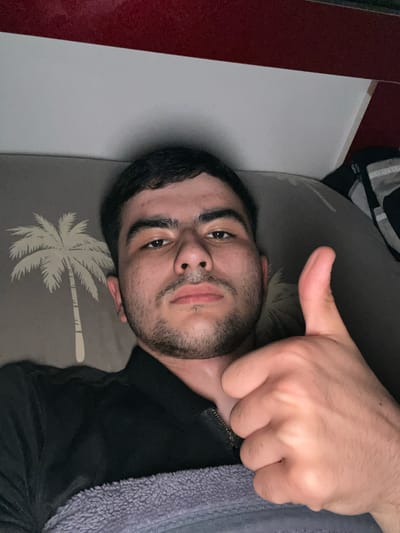 A person is lying down, facing the camera, and giving a thumbs-up. They appear to be on a bed or possibly a couch, as indicated by the patterned pillow or cushion beneath their head. The pillow/cushion features a palm tree design. The person has dark hair, eyebrows, and a beard. Part of what seems to be a dark-colored garment is visible, along with a gray fuzzy material, possibly a blanket or robe. There is a white surface next to the pillow with a dark red piece on the left of the white surface. The location is unknown.