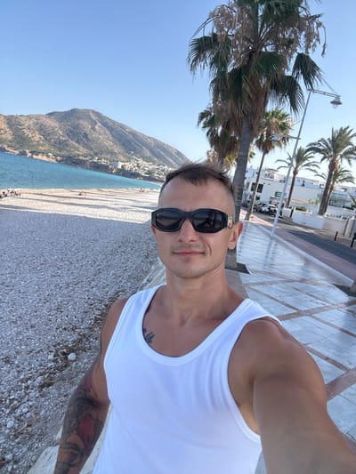 A man wearing a white tank top and black sunglasses takes a selfie on a beach promenade in Altea, Spain. He is standing on a stone pathway with a pebble beach and the Mediterranean Sea to his left. Palm trees and white buildings line the promenade behind him. In the background, mountains rise behind the coastal town. Several people are visible relaxing on the beach. The sky is clear and blue.