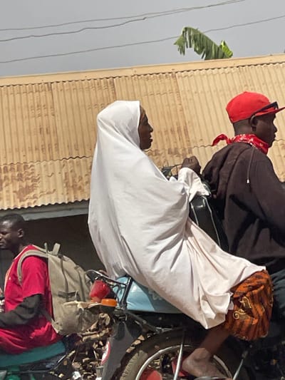 The scene depicts an urban street in Unguwar Gabas, Nigeria. In the foreground and mid-ground, three individuals are seen in various states of transit on motorcycles. An adult female, dressed in a long, flowing white garment covering her head and body, and an orange and brown patterned lower garment, rides side-saddle as a passenger on a motorcycle, facing left. Her bare feet are visible near the rear wheel. The male operator of this motorcycle, positioned in front of her, is seen in profile wearing a dark brown long-sleeved shirt, a red baseball cap worn backward, and a red patterned bandana around his neck. To the left, a second adult male, on a separate motorcycle, wears a red short-sleeved shirt over a dark long-sleeved shirt, and a light-colored backpack. The visible portion of the motorcycle carrying the female passenger includes a blue seat, red taillights, and orange indicators. The background features a weathered, light brown corrugated metal structure, likely a building or fence. Above this structure, a light grey sky shows thin overhead utility wires and a cluster of large green leaves, consistent with banana foliage, in the upper right.