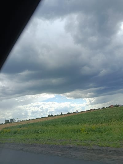 The image depicts a rural landscape with a focus on the sky and fields. The sky is filled with dramatic, dark clouds, suggesting an impending storm or overcast weather. Below the clouds, there are patches of lighter sky and scattered white clouds. The foreground shows a green field that appears to be cultivated, with rows of plants visible. In the distance, there are hints of buildings, possibly a village or houses, nestled on a slight incline. A prominent blue and yellow structure, likely a church with a dome, is visible among the buildings on the horizon. The overall scene conveys a sense of an open, natural environment under a dynamic sky. The location is identified as Biloziria, Ukraine.