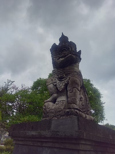 A stone statue of a mythical creature stands against a cloudy sky in Gianyar, Indonesia. The statue is intricately carved, depicting a fearsome figure with a detailed headdress, ornate jewelry, and muscular limbs. It is positioned on a stone pedestal, which is part of a larger stone structure. Lush green trees with some purple flowers frame the background, adding a touch of nature to the scene. The lighting suggests an overcast day.