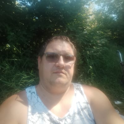 A man wearing sunglasses and a white tank top is pictured outdoors in a natural environment. He is seated with tall green grass and foliage behind him. The lighting suggests it is daytime, with sunlight filtering through the leaves. The man's face is directed towards the camera, and he appears to be looking directly at it. The overall impression is a casual, candid snapshot taken in a park or natural setting. The location is not discernible from the image itself.