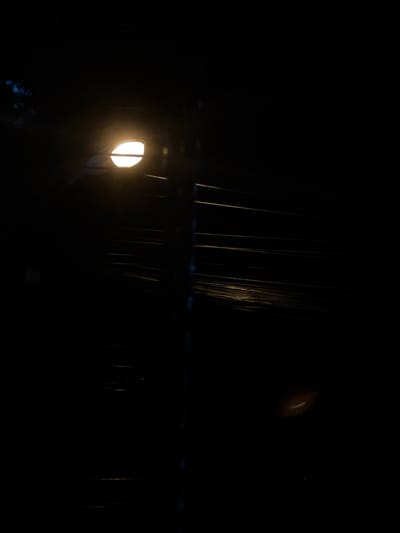In Kathu, Thailand, at night, a streetlamp illuminates a dark scene. The light from the lamp, a bright white oval, casts a glow on the surrounding darkness. A vertical pole, likely part of the streetlamp assembly, is visible in the center of the frame, running from top to bottom. A network of thin, horizontal wires stretches across the background, catching some of the light and appearing as faint yellow lines. To the right of the pole, a blurry, oval-shaped light artifact, possibly a lens flare, is visible in the dark space. The overall impression is one of a quiet, dimly lit urban environment at night.