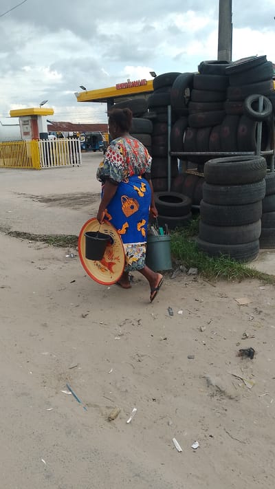 A woman in a colorful dress and blue wrapper walks across a sandy, debris-strewn street in front of a gas station and a stack of tires. She carries a round metal tray with a black bucket hanging from it in her left hand, and a green container with various items in her right hand. The gas station sign reads "PANews GLOBAL OIL". The sky is overcast. The scene appears to be in Agbarho, Nigeria, based on the provided location context.