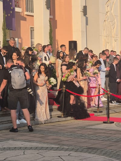 A group of people dressed in formal attire stand along a red carpet, lined by stanchions with a red rope. They are arranged on a grey stone tiled surface near a light-colored building. Some people are holding bouquets. A photographer is present, taking pictures with a professional camera. An EU flag is visible in the background. The event is located in Pavlikeni, Bulgaria.