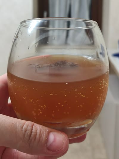 A person's hand holds a clear glass containing a carbonated amber liquid. The liquid has small bubbles visible throughout. There are water droplets on the exterior of the glass. The background is blurred, suggesting an indoor environment. The image does not provide information about the city of Campochiaro, Italy.