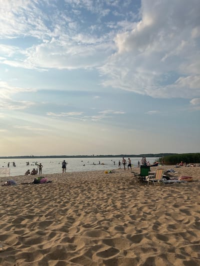 The image depicts a sandy beach adjacent to a body of water, likely a lake or river, under a partially cloudy sky. The beach is populated with numerous individuals engaged in various activities. In the foreground, the sand is textured with footprints and ripples. Towards the middle ground, people are seen wading and swimming in the water. Some individuals are sitting or lying on the sand, some on beach towels and blankets. Two beach chairs are set up on the sand. In the distance, a line of trees forms the horizon. The overall scene suggests a recreational setting on a warm day. The provided hints suggest the location is within the Загародскі сельскі Савет, Belarus.