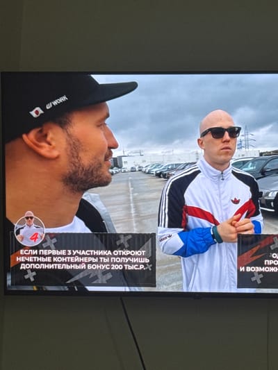 The image shows a television screen displaying a scene with two men. The man on the left is wearing a black baseball cap with a Japanese flag and white text that appears to read "WORK". He has a beard and is in profile, looking to the right. The man on the right is bald, wearing sunglasses, and a white and blue tracksuit with red stripes. He is looking forward. Both men appear to be outdoors, possibly in a parking lot, with a line of cars in the background. There is a graphic overlay on the bottom of the screen with Russian text and images. The scene does not depict any recognizable landmarks of Neubrandenburg, Germany.