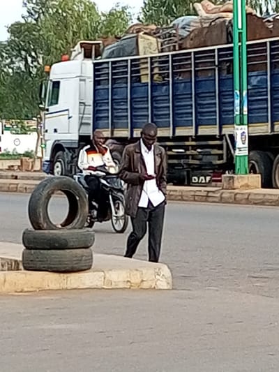 In Tumfure, Nigeria, on a paved street, a man in a brown jacket and white shirt walks past a stacked pile of three tires. Behind him, another man rides a motorcycle, also wearing a white jacket with stripes and black pants. A large blue truck, loaded with cargo and appearing to be a DAF model, is parked on the right side of the road. The truck has a white cab with an orange hazard light on top. The environment is characterized by trees and a sidewalk with a stone border.