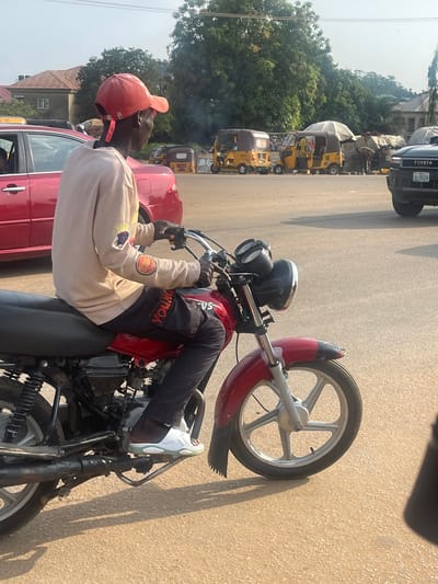 A young Black man wearing a red baseball cap, a light-colored hooded sweatshirt with patches on the sleeves, and dark pants is sitting on a red motorcycle. His right foot is resting on the motorcycle's footpeg, and he is wearing white sneakers. The man appears to be looking off to the side, away from the camera, with a neutral expression.
The motorcycle is parked next to a red car. In the background, there are several yellow auto-rickshaws, also known as "keke napep," and a black Toyota SUV. Trees and buildings are visible further in the background, suggesting an urban or semi-urban environment. The scene takes place on a paved road under a bright sky, indicating it is daytime. The lighting suggests it could be mid-morning or afternoon.
The overall impression is a candid snapshot of everyday life in a Nigerian village or town. There is no specific event or activity clearly depicted beyond the man being on his motorcycle. The presence of multiple auto-rickshaws points to a location where this is a common mode of transportation. Visible text includes "TOYOTA" on the SUV and "YOUNG" on the man's jacket. There are also some patches with text on the man's sweatshirt, but they are not clearly legible.