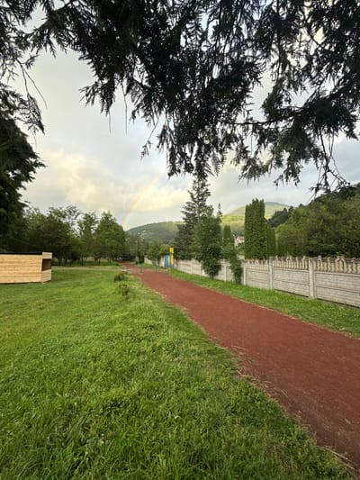 This is a vertical, wide-angle photograph taken outdoors on an overcast day. The image features a lush green landscape with a red, dusty running track on the right side. In the foreground, the branches of dark green coniferous trees frame the left side of the shot.

The background reveals a gentle slope covered with trees and a light-colored, decorative stone fence. A faint rainbow arcs across the cloudy sky, suggesting recent or ongoing rain. Further in the distance, a white house with a red roof is visible nestled amongst the greenery on the hillside. At the bottom right of the image, a simple wooden structure, possibly a bench or a small shed, is present. The scene evokes a sense of tranquility and natural beauty, possibly in a park or recreational area. The time of day appears to be late afternoon or early evening, given the soft, diffused light. The location is identified as Dilijan, Armenia.