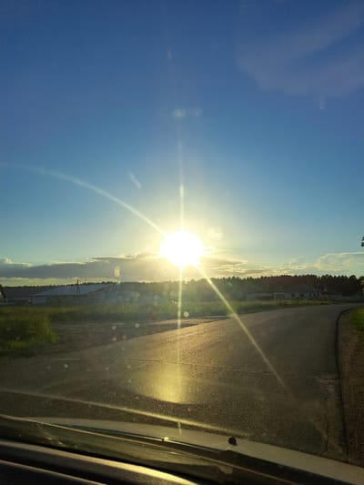 This image captures a road winding through a rural landscape under a bright, sunlit sky. The sun is positioned high and to the left of center, emitting strong rays and creating lens flare effects on the camera. The road is paved and appears to be in good condition, with a light-colored line marking its center. To the left of the road, there is a grassy field with some sparse vegetation. Beyond the field, a line of trees forms a dark silhouette against the horizon. In the distance, there are several buildings, including what appear to be farm structures or warehouses, one with a distinct white roof. The sky is a clear, vibrant blue with a few wispy clouds on the horizon, catching the sunlight. The foreground of the image shows the hood and windshield of a vehicle, suggesting the photo was taken from inside a car. The overall scene depicts a sunny day in a rural setting.