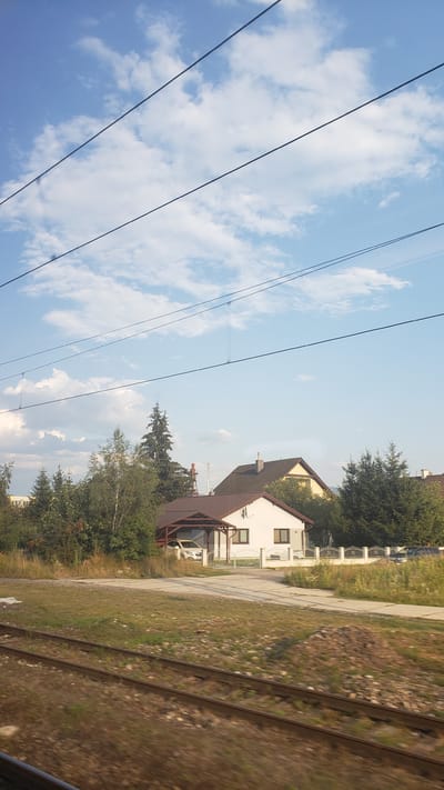 The image depicts a view from a moving train, as indicated by the blurred foreground of railroad tracks and the presence of overhead wires. The scene is set in Liptovská Teplá, Slovakia, under a partly cloudy sky. In the mid-ground, several residential houses are visible, nestled amongst trees and vegetation. One house in particular is a white structure with a brown roof and a covered carport sheltering a white car. Adjacent to it is another house with a yellow facade and a darker brown roof. A white fence encloses a portion of the property. The surrounding area features a mix of greenery, including trees and grassy areas, with a pile of dirt or debris in the foreground. In the background, a tall industrial structure with a red top is visible, suggesting proximity to an industrial zone or a local landmark. The overall impression is a glimpse of a suburban or semi-rural landscape.