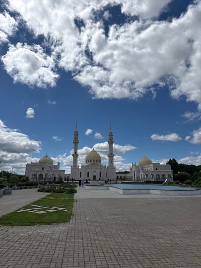 A vertical, wide shot shows the White Mosque in Bulgar, Russia, under a bright blue sky dotted with fluffy white clouds. The mosque's white facade, golden domes, and tall minarets are visible. The building is situated next to a pool of clear blue water, reflecting the sky. A paved walkway made of grey bricks runs alongside the pool, with a strip of green grass featuring a diamond pattern of white stone inlays. Several people are walking on the walkway and near the mosque, suggesting a daytime visit. The weather appears pleasant, with ample sunlight. The overall scene evokes a sense of peace and architectural grandeur.