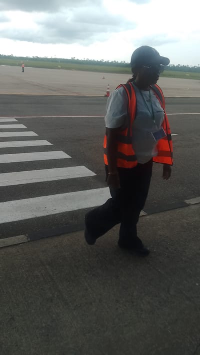 A woman wearing a dark baseball cap, sunglasses, and an orange high-visibility vest over a light blue shirt walks across a pedestrian crossing at an airport. She is also wearing dark pants and dark shoes. The pedestrian crossing has thick white stripes. In the background, an empty runway is visible, along with orange traffic cones, and distant greenery and trees under a cloudy sky. The woman is the only person clearly depicted in the foreground. The scene appears to be at the airport in Orerokpe, Nigeria, given the context.
