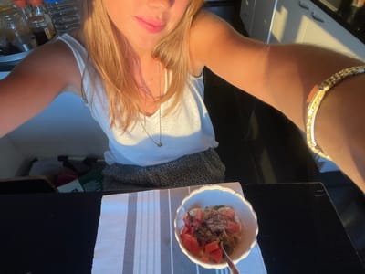 A partial view of an individual, identified as being in Aldoar, Foz do Douro e Nevogilde, Portugal, is captured in a selfie-style photograph. The subject, a person with light-colored hair, is visible from the upper chest to the top of the head, wearing a white sleeveless top, a delicate necklace with a small pendant, and a gold-toned bracelet on the right wrist. A leopard-print patterned lower garment is also visible. The person is seated at a dark-surfaced table featuring a white and grey striped placemat. On the placemat, a white scalloped bowl contains a meal consisting of a light base, topped with diced red fruit, dark seeds, and green herbs, with a spoon placed inside. The background suggests an indoor domestic setting, likely a kitchen or dining area, showing glass bottles and jars on a counter to the left and white cabinetry with a dark handle to the right. Strong natural light, originating from the right, illuminates the scene, creating distinct highlights and shadows.