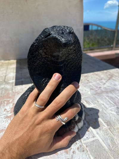 A person's hand, adorned with two silver rings, is partially obscuring a black, textured sculpture of a cobra's head. The sculpture exhibits a scaled pattern and appears to be made of a dark, possibly lacquered material. The hand is positioned in front of the cobra's head, with fingers gently touching the textured surface. The scene is set outdoors, on a weathered, light-colored wooden table. In the background, a bright blue sky with a few white clouds is visible, along with the shimmering expanse of the ocean, suggesting a coastal location. A blurred railing with dark posts and glass panels separates the immediate foreground from the distant seascape, implying a balcony or terrace setting. The overall lighting suggests a sunny day. The specific location in Ko Samui, Thailand, is not discernible from the visual information alone.