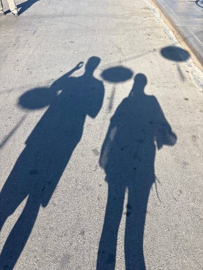 Two shadows of people are cast on an asphalt surface. The person on the left is taller, with their shadow extending longer across the ground. They appear to be holding something in their raised hand, casting a circular shadow on the ground. The person on the right is shorter, with their shadow also on the asphalt. The surface shows some wear and tear, with small dark spots and cracks visible. In the background, a concrete curb and sidewalk are visible, with some tiles laid out. The scene suggests a sunny day, with the shadows clearly defined. The specific location is Hội An Ward, Vietnam.