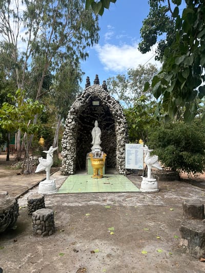 An outdoor religious shrine, possibly Buddhist or Taoist, is depicted in a natural setting under a bright, cloudy sky in Cam Ranh, Vietnam. The central feature is a grotto-like structure covered entirely with numerous small, light-colored shells or pebbles, giving it a unique textured appearance.
Inside the grotto, a white, standing statue of a deity, likely Guanyin (Bodhisattva of Compassion), is prominently placed on a white pedestal. To the right of the statue (viewer's left), a golden, ornate censer or offering bowl rests on a green-tiled platform that extends outwards from the grotto. Flanking the shrine on either side of the platform are white statues of stylized birds, possibly cranes or phoenixes, perched on pedestals.
The ground in the immediate foreground is a concrete or paved area, scattered with a few green leaves. Further to the right, two cylindrical stone pedestals, also textured with small stones, are visible. The shrine is surrounded by lush green trees and foliage, with tall, slender-trunked trees providing a natural canopy and backdrop. The sky above is a clear blue with scattered white, fluffy clouds, indicating daytime, likely morning or early afternoon.
To the right of the grotto, a white sign with black text on a pole displays what appears to be a list, possibly names or dedications, though the specific text is unreadable due to distance. No people are visible, and no specific event is taking place, contributing to a serene and tranquil atmosphere at this spiritual site.
