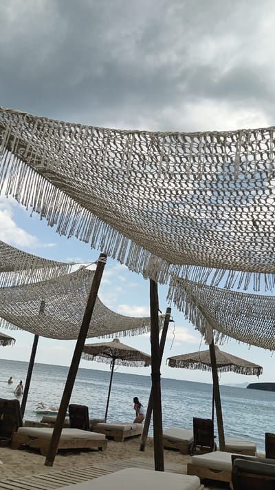 A beach resort in Nea Peramos, Greece, features several rustic, open-air cabanas with woven canopies and fringed edges, supported by wooden poles. Underneath these canopies are rows of lounge chairs, many of which are unoccupied. The beach itself is sandy, with wooden platforms laid out in some areas. The sea is visible in the background, with a few people swimming or wading in the water. One person is floating on a white inflatable raft, while another is sitting on a lounge chair near the water's edge. The sky is overcast with dramatic clouds.