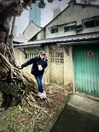 A medium shot captures a woman wearing a dark jacket, blue jeans, and a blue baseball cap posing next to a large tree with exposed roots. She holds a pink cup in her right hand and leans her left arm on the tree. The background features a weathered building with a green door and a diamond-shaped red decoration with Chinese calligraphy. In the distance, a tall, modern skyscraper is visible against a cloudy sky. The scene appears to be located in an older district of Taipei, Taiwan, contrasting the traditional architecture with the modern urban landscape.