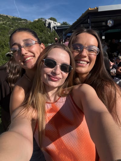 In a selfie taken outdoors, three young women are posing together under a bright blue sky with some scattered clouds. The woman on the left, with dark hair pulled back, wears clear-rimmed glasses and smiles with her eyes mostly closed. She has a dimple on her right cheek. Next to her, the woman in the center, with blonde hair and sunglasses, smiles widely, revealing her teeth. She is wearing an orange and white patterned tank top. The woman on the right, with long brown hair and clear-rimmed glasses, also smiles broadly, her mouth open. She is wearing a dark top. In the background, a rocky, tree-covered hillside is visible, with a building structure with a dark roof and yellow accents overhead. Several other people are present in the background, some standing and some seated at tables, suggesting a gathering or outdoor dining area. The overall impression is of a casual, happy moment shared by friends. The location is identified as Špiljari, Montenegro.