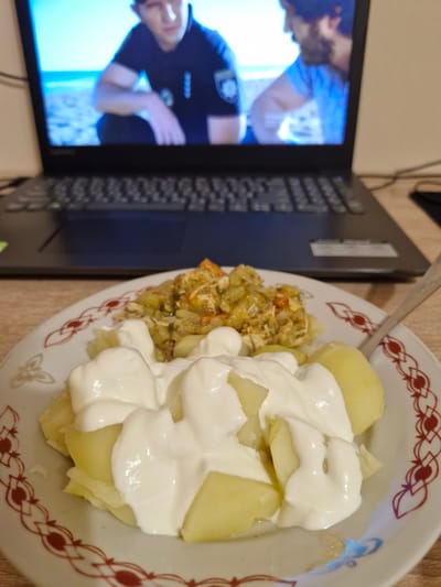 A close-up, slightly blurry, eye-level shot shows a plate of food in the foreground and a laptop in the background. The plate, adorned with a red and gold patterned rim, contains boiled potatoes topped with a generous dollop of sour cream. Behind the plate, a laptop displays a scene from a television show or movie, featuring two men on what appears to be a beach. The laptop is open and positioned on a wooden surface, likely a desk or table. The overall impression is of a casual meal enjoyed while watching content on a screen. There is no indication of the specific location in Lviv, Ukraine.