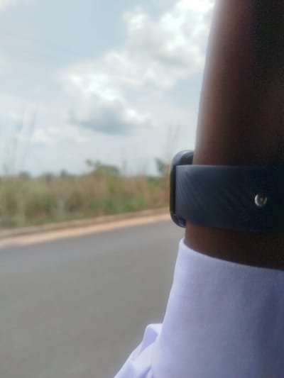 The image shows a close-up of a person's arm wearing a black fitness tracker on the left wrist. The person is wearing a white collared shirt. The background is blurred, showing a paved road, grassy vegetation, and a cloudy sky. The location is Ukwulu, Nigeria.