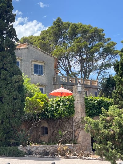 A medium shot captures a traditional stone and stucco house, partially obscured by lush greenery and tall trees, under a bright blue sky with scattered white clouds. The house features a terracotta tiled roof and several windows with blue shutters. A balcony with ornate railings is visible on an upper level. In the foreground, a vibrant red umbrella sits atop a dense wall of ivy. A stone pillar with a lantern stands to the right of the umbrella. The lower part of the image reveals a rock wall bordering a garden with various plants, including succulents and dry brush. The scene is situated in the town of Pollença, Spain.